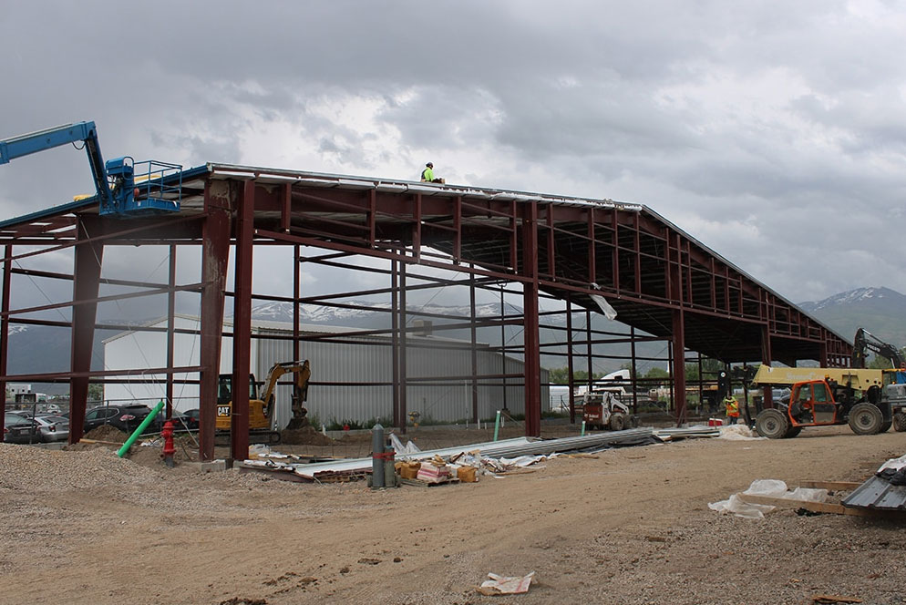 Industrial construction site with a large steel-framed building under development, featuring contractors on an aerial lift installing the roof and heavy machinery operating below.